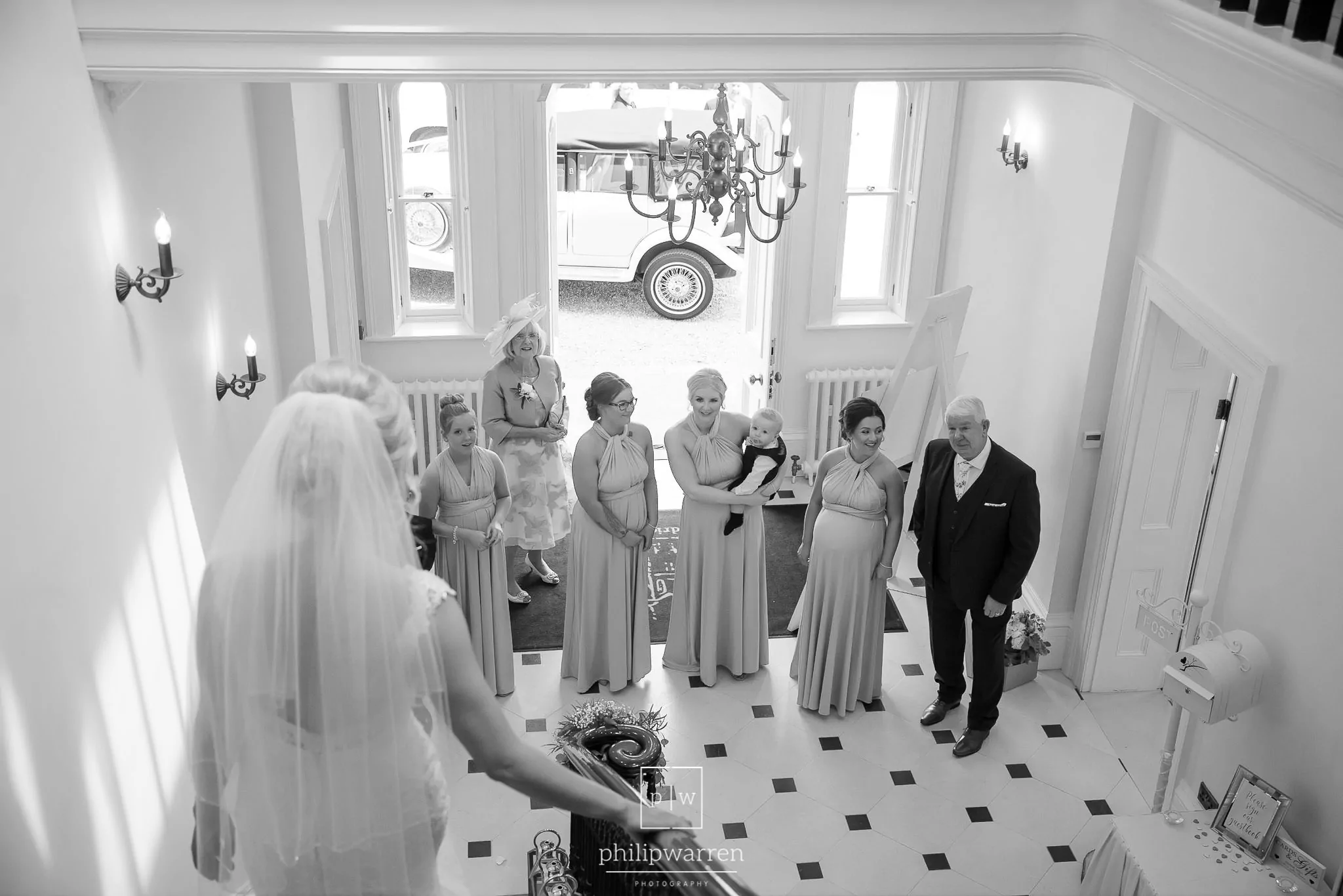 black and white photo taken from behind the bride with her mother and father and bridesmaids waitingat at he bottom of the stairs at this welsh wedding venue