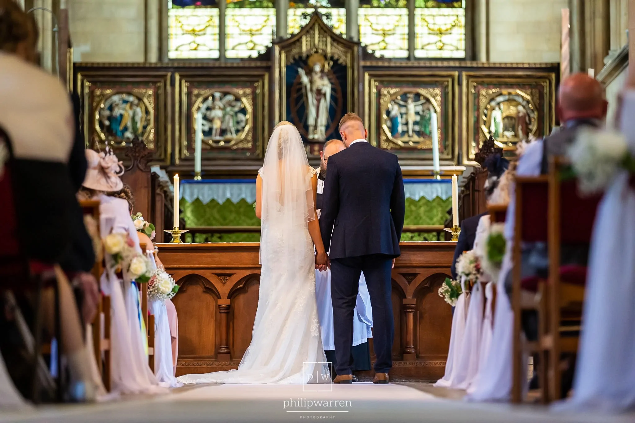 photo taken from at the back of the church lookign down the aisle at the bride and groom holding hands during the ceremony at st marys church in chepstow